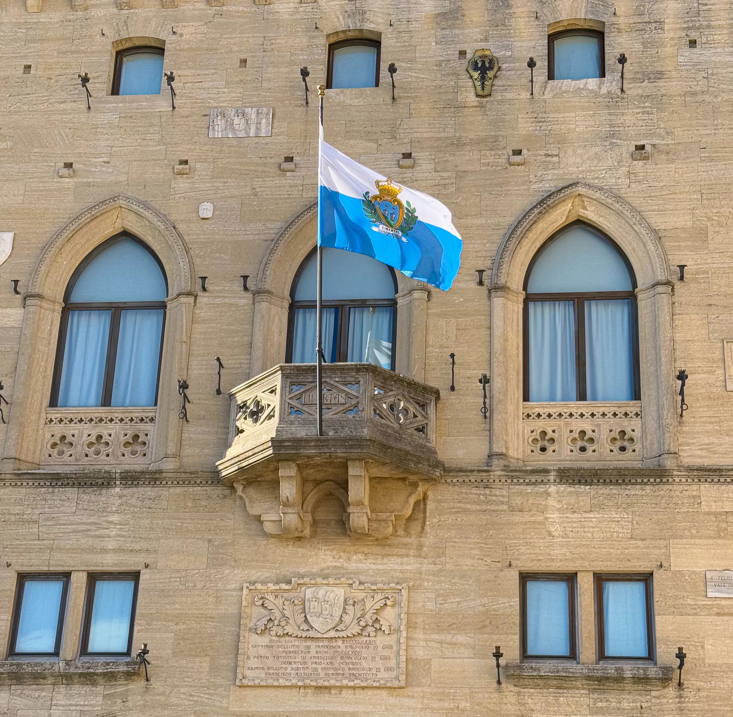 Флаг Сан-Марино над резным каменным балконом Палаццо Пубблико со стрельчатыми окнами в ярком солнечном свете.