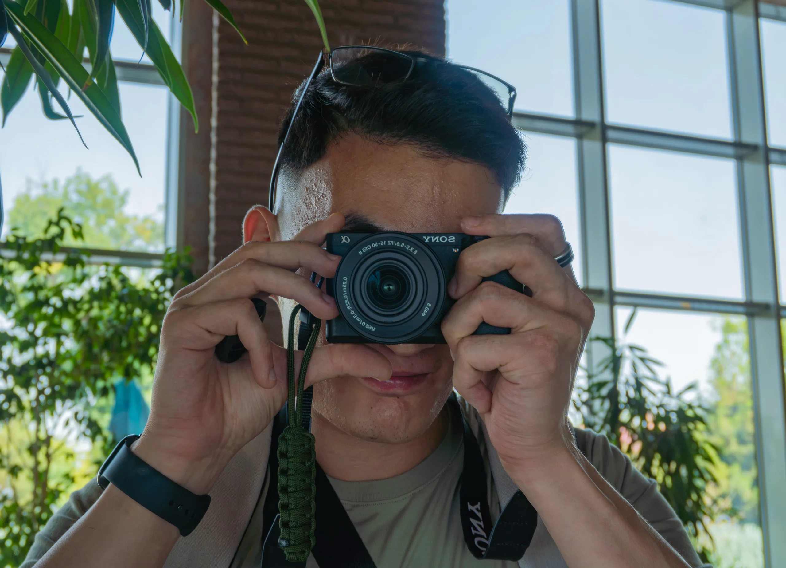 Автопортрет фотографа в зеркале кафе: камера Sony у лица, за спиной большие окна и зелень в дневном свете.