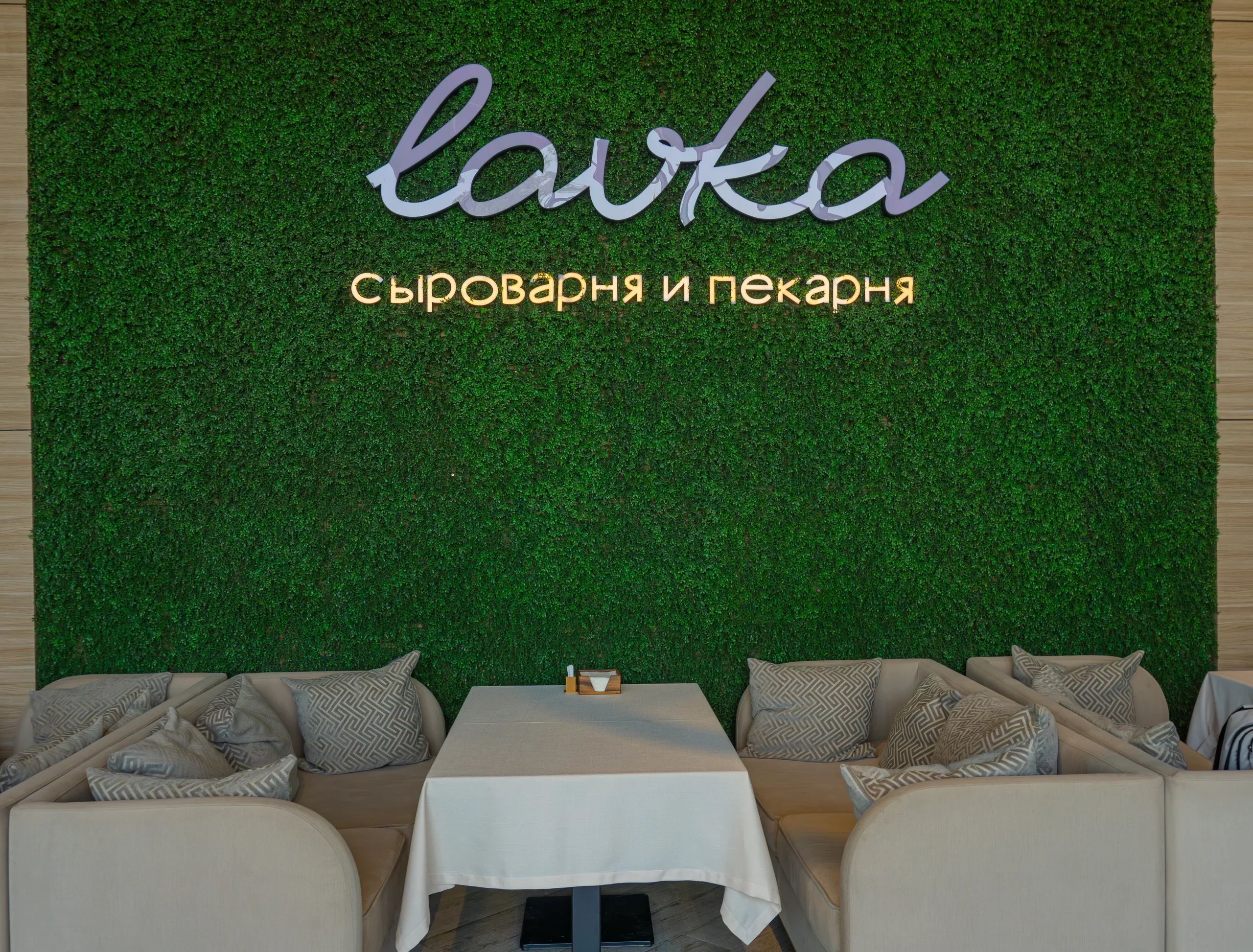 Стена из зелени с вывеской Lavka, под ней светлые диваны с подушками и стол с белой скатертью при дневном свете.