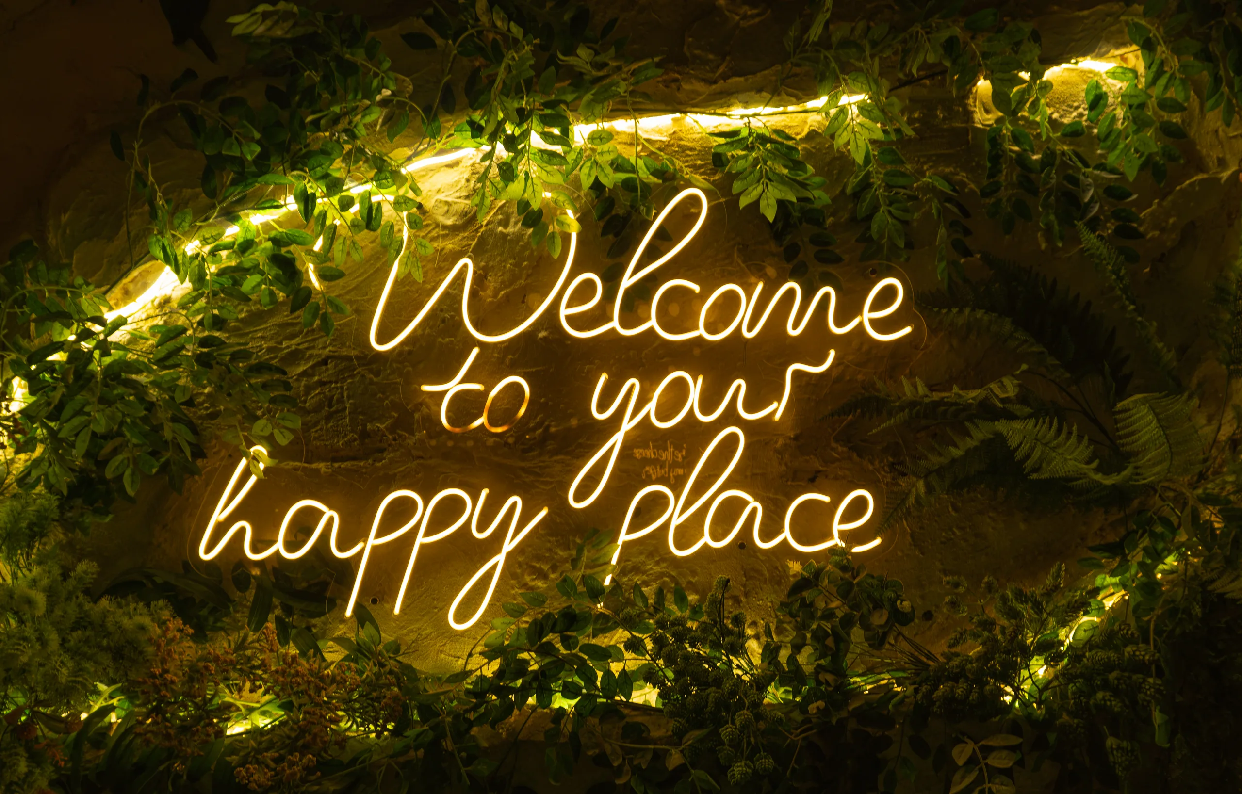Неоновая надпись Welcome to your happy place тёплого жёлтого свечения на стене среди листвы.