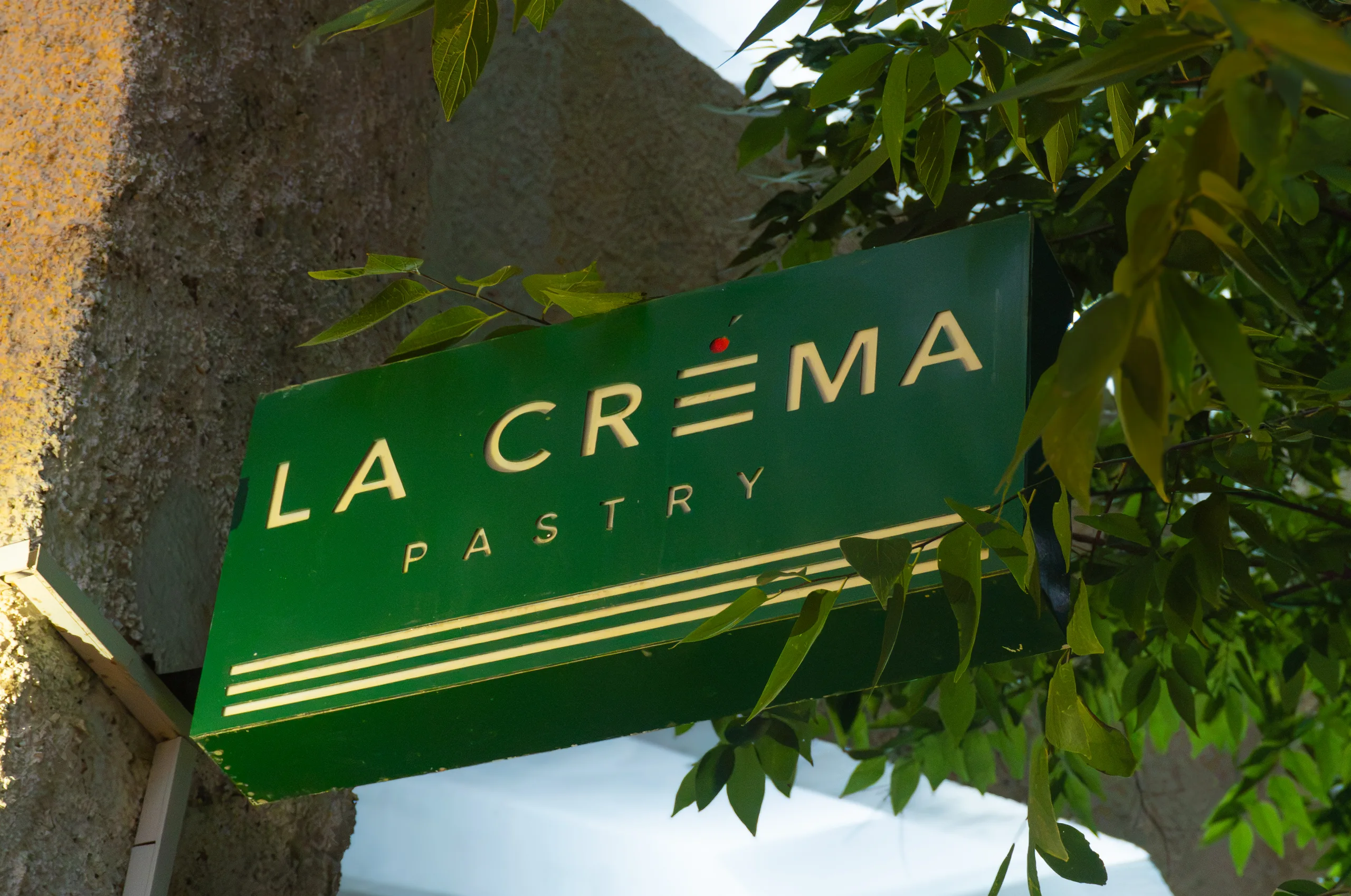Зелёная вывеска La Créma Pastry с кремовыми буквами среди листвы на углу здания при дневном свете.