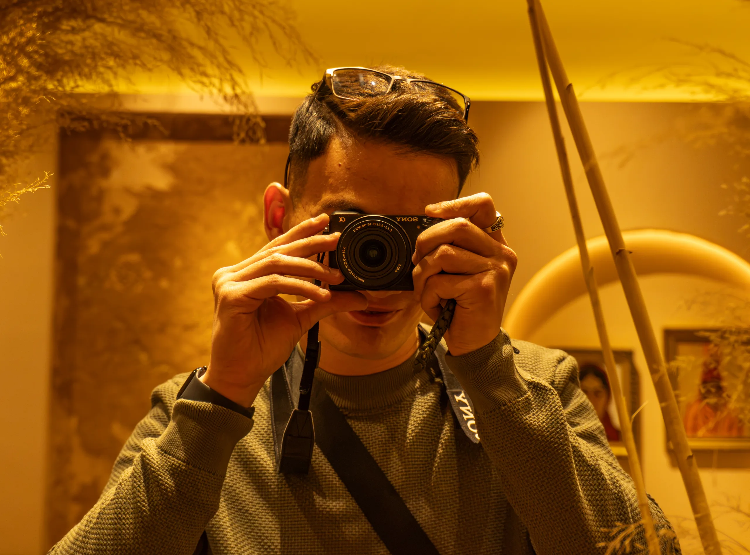 Автопортрет фотографа в зеркале кафе TOR: камера Sony у лица, янтарный свет и сухие травы по краям кадра.