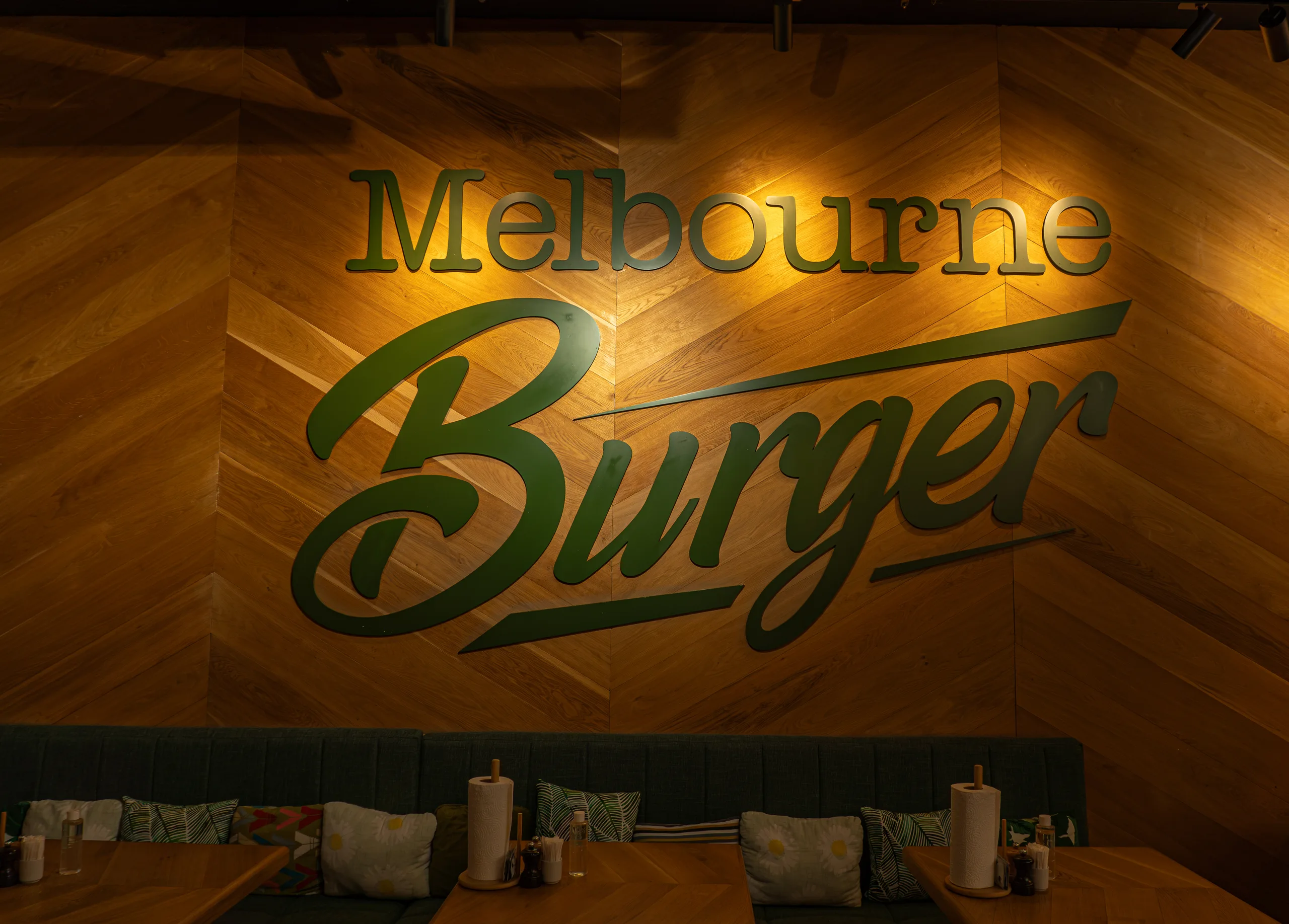 Зелёная объёмная вывеска Melbourne Burger на стене из деревянных панелей ёлочкой над серым диваном, тёплая подсветка.