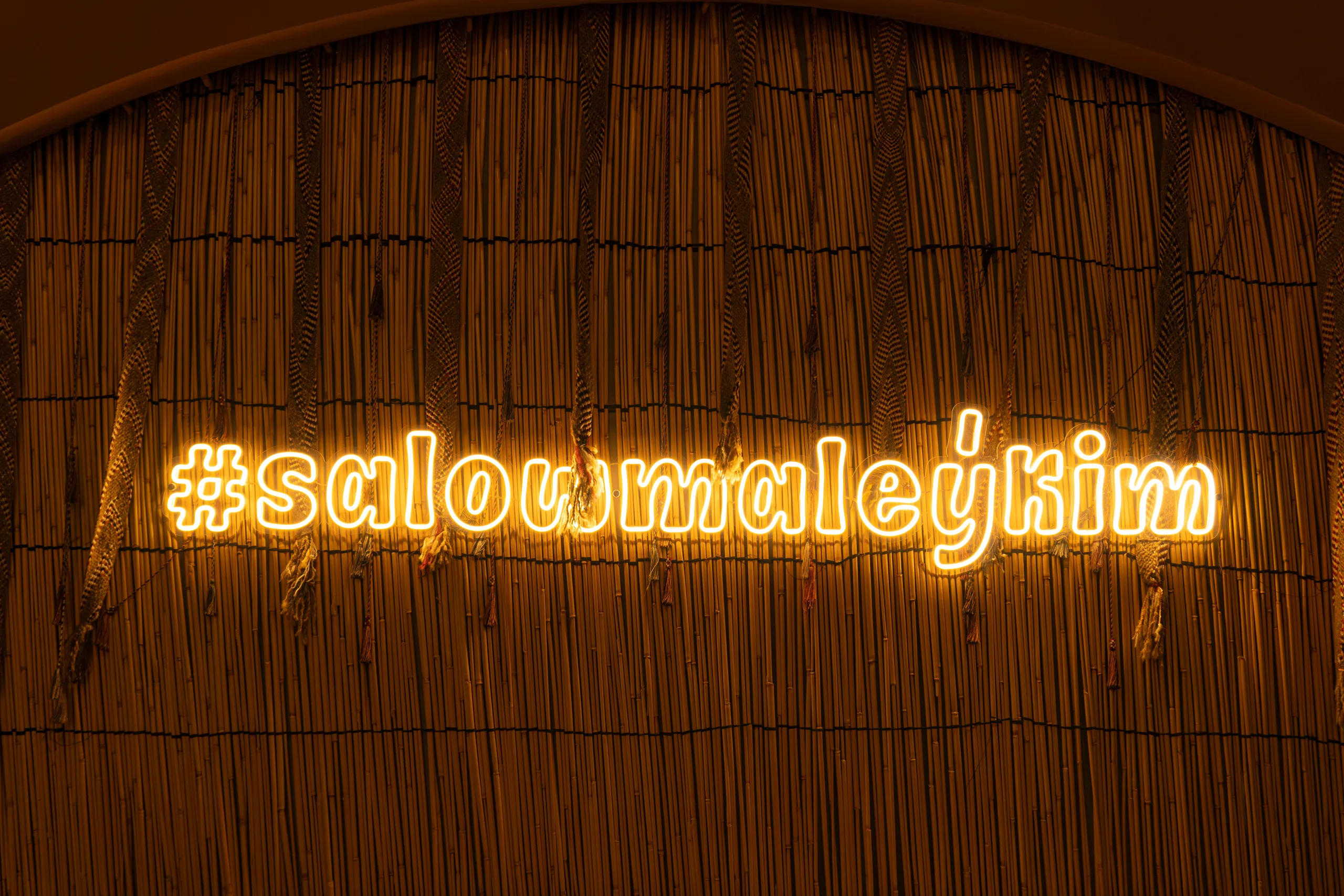 Жёлтая неоновая надпись #salowmaleýkim на фоне плетёной камышовой стены в тёмном зале.