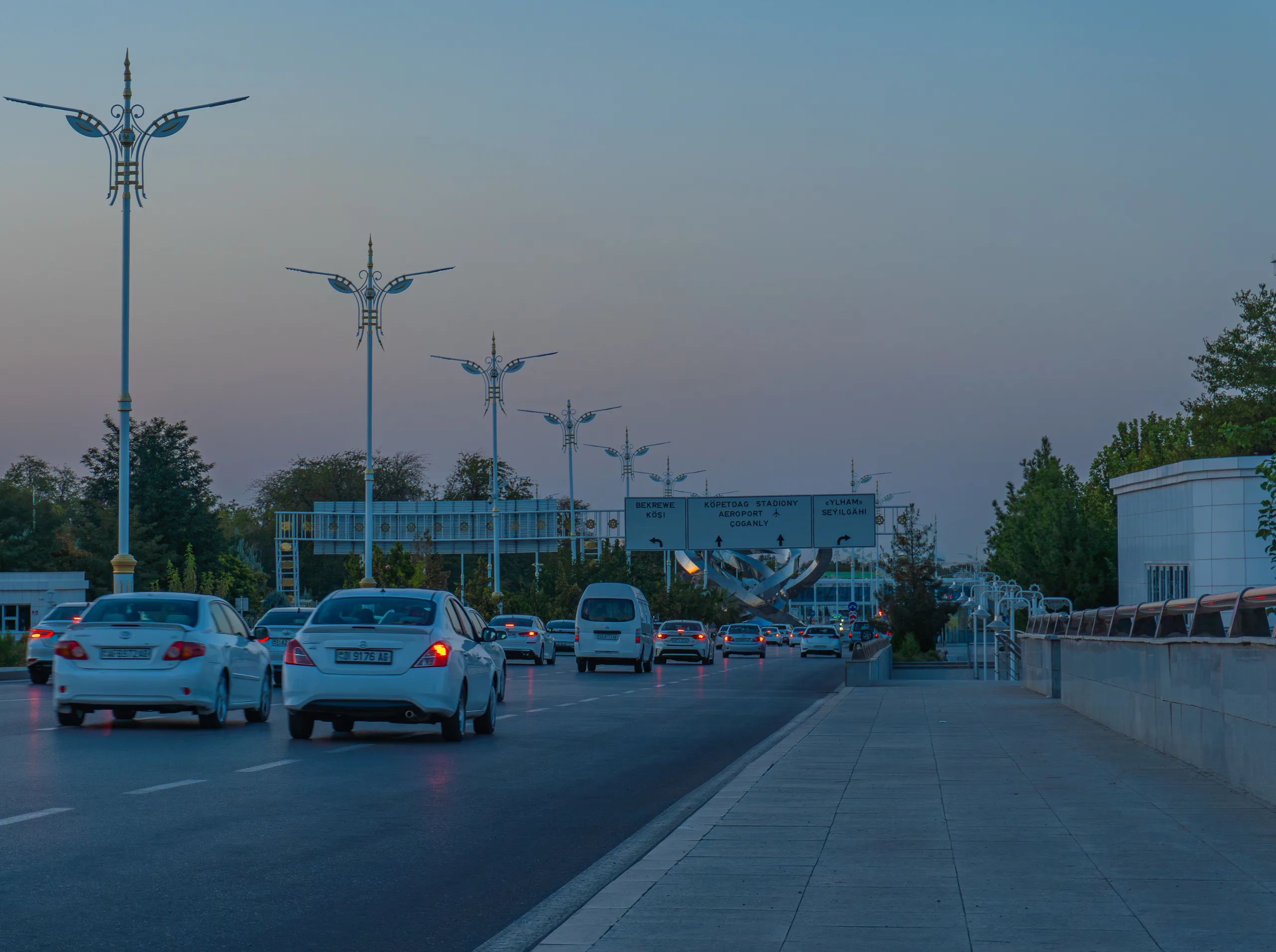 Поток машин на проспекте Ашхабада в сумерках, белые декоративные фонари и дорожные указатели на туркменском языке.