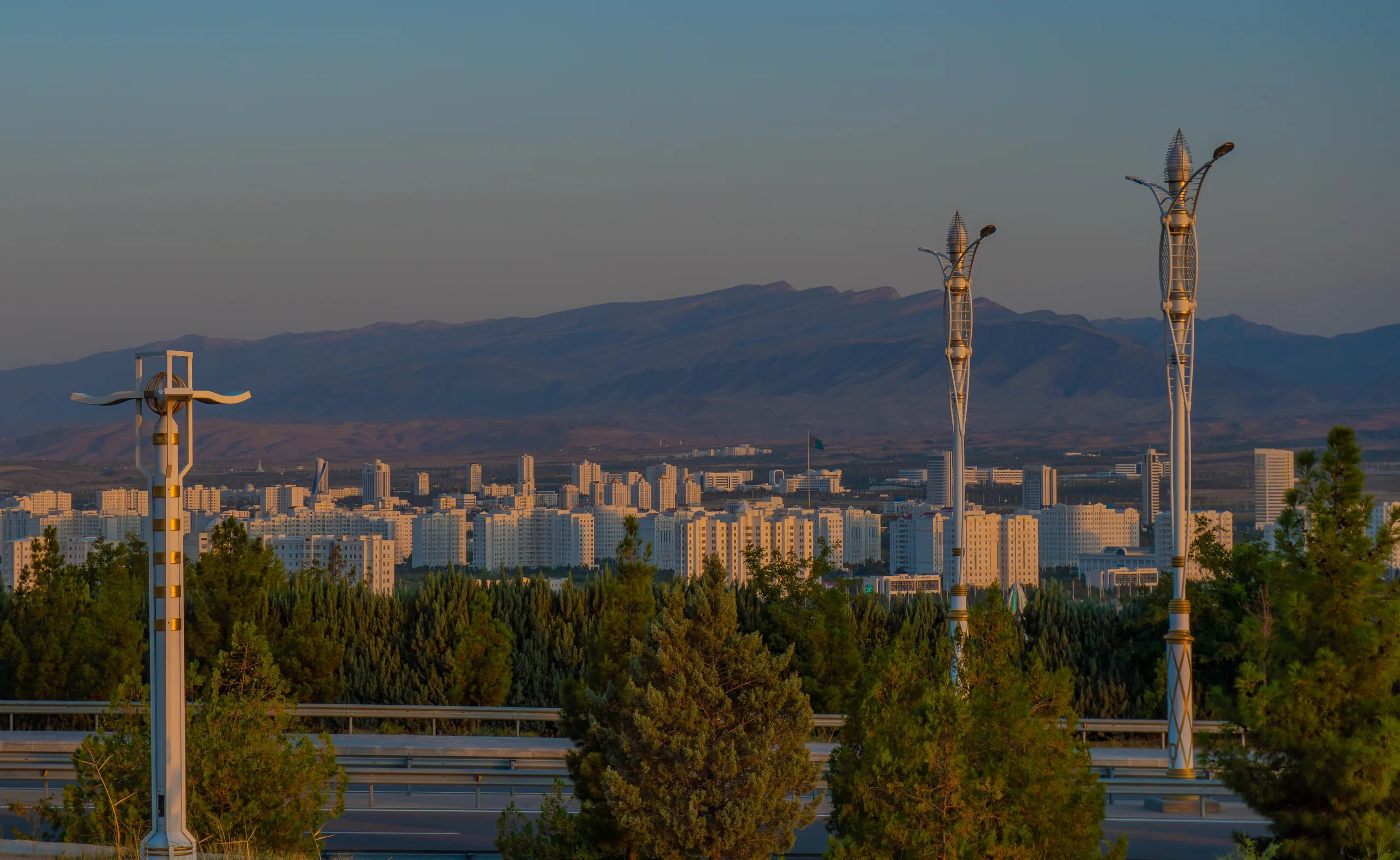Панорама белого города на фоне гор в тёплом вечернем свете, на переднем плане два высоких декоративных фонаря и хвойные деревья.
