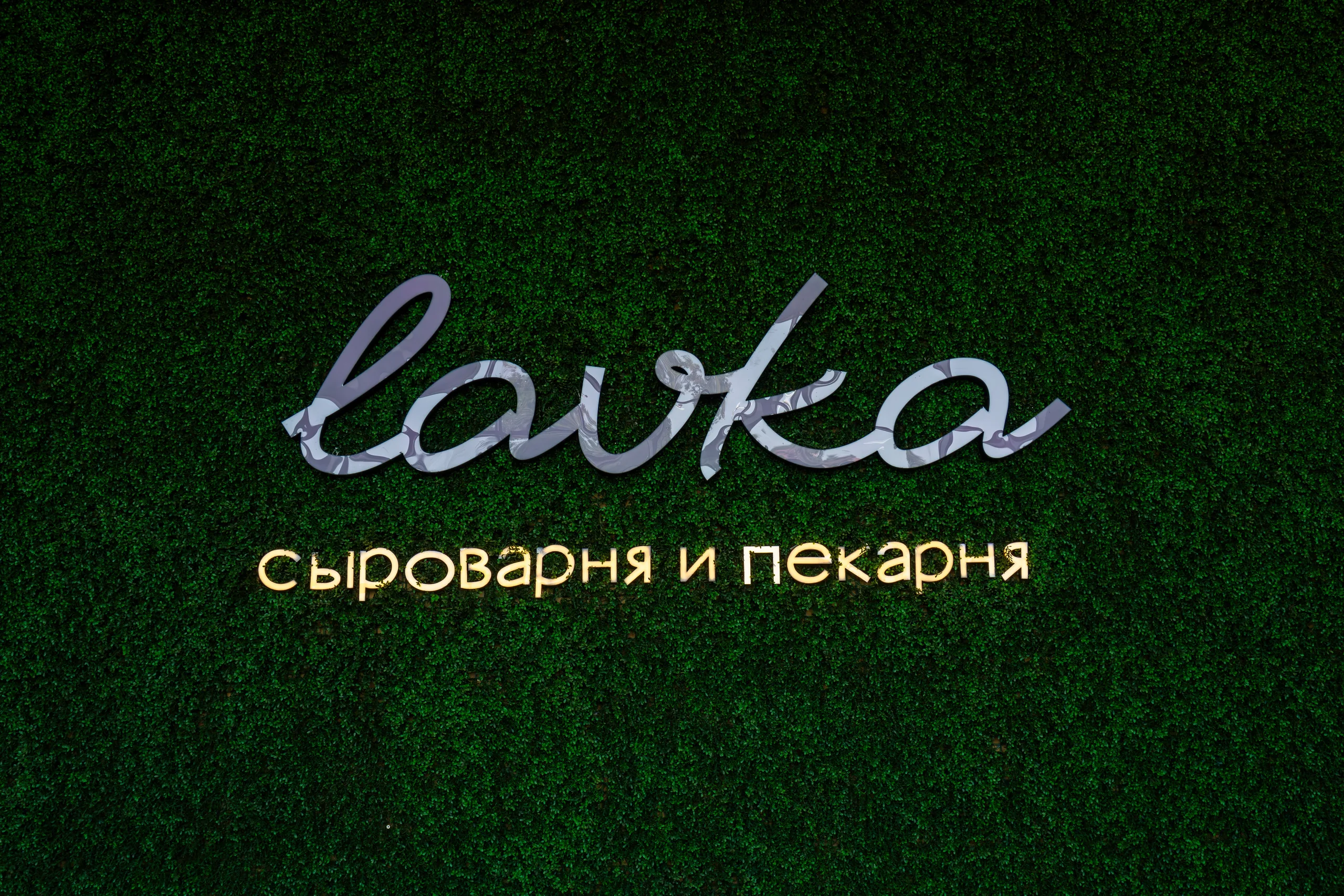 Вывеска Lavka с подсвеченной подписью «сыроварня и пекарня» на тёмной стене из искусственной зелени.