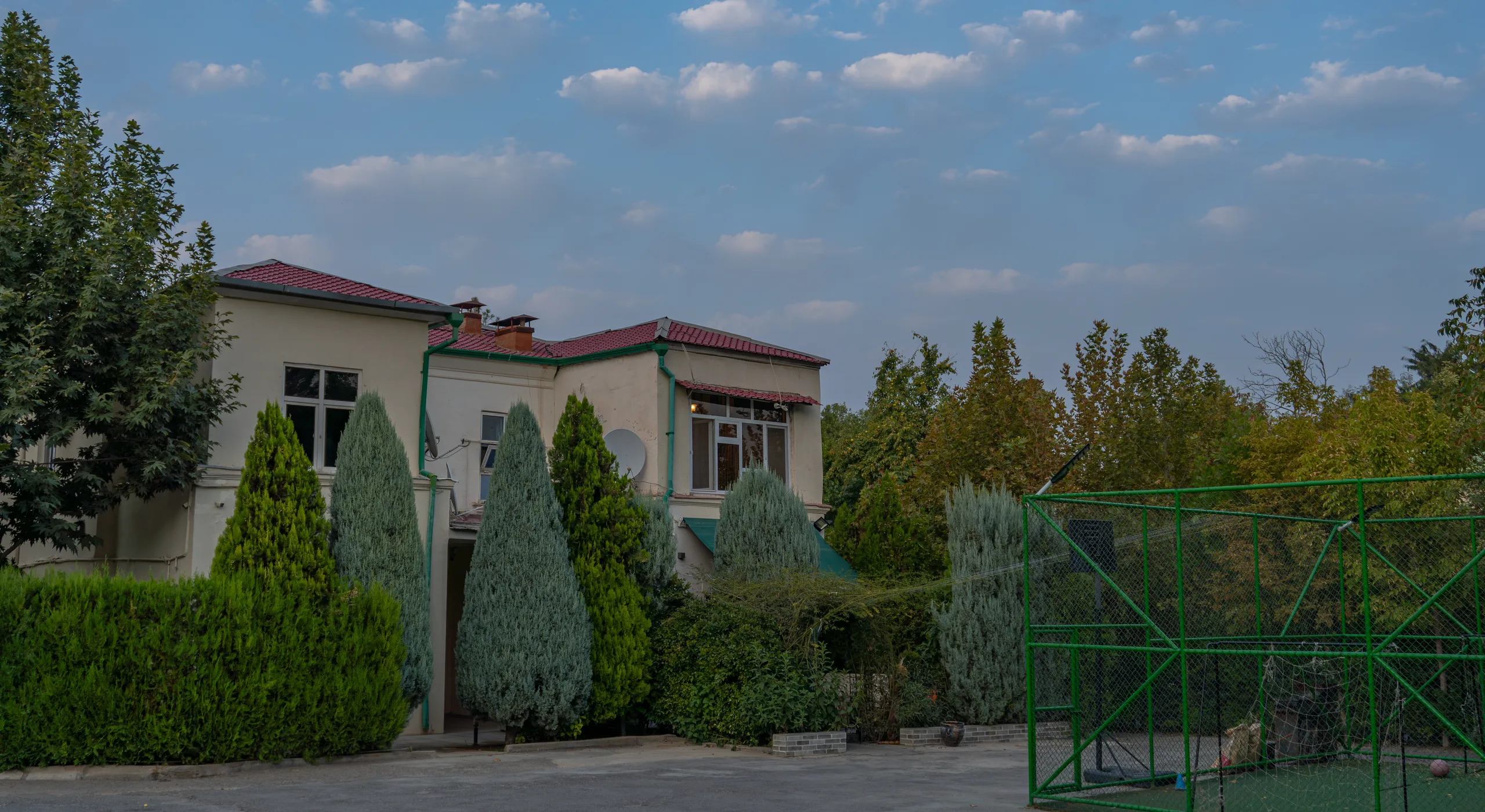 Двухэтажный дом с красной крышей за кипарисами и живой изгородью, рядом спортивная площадка.